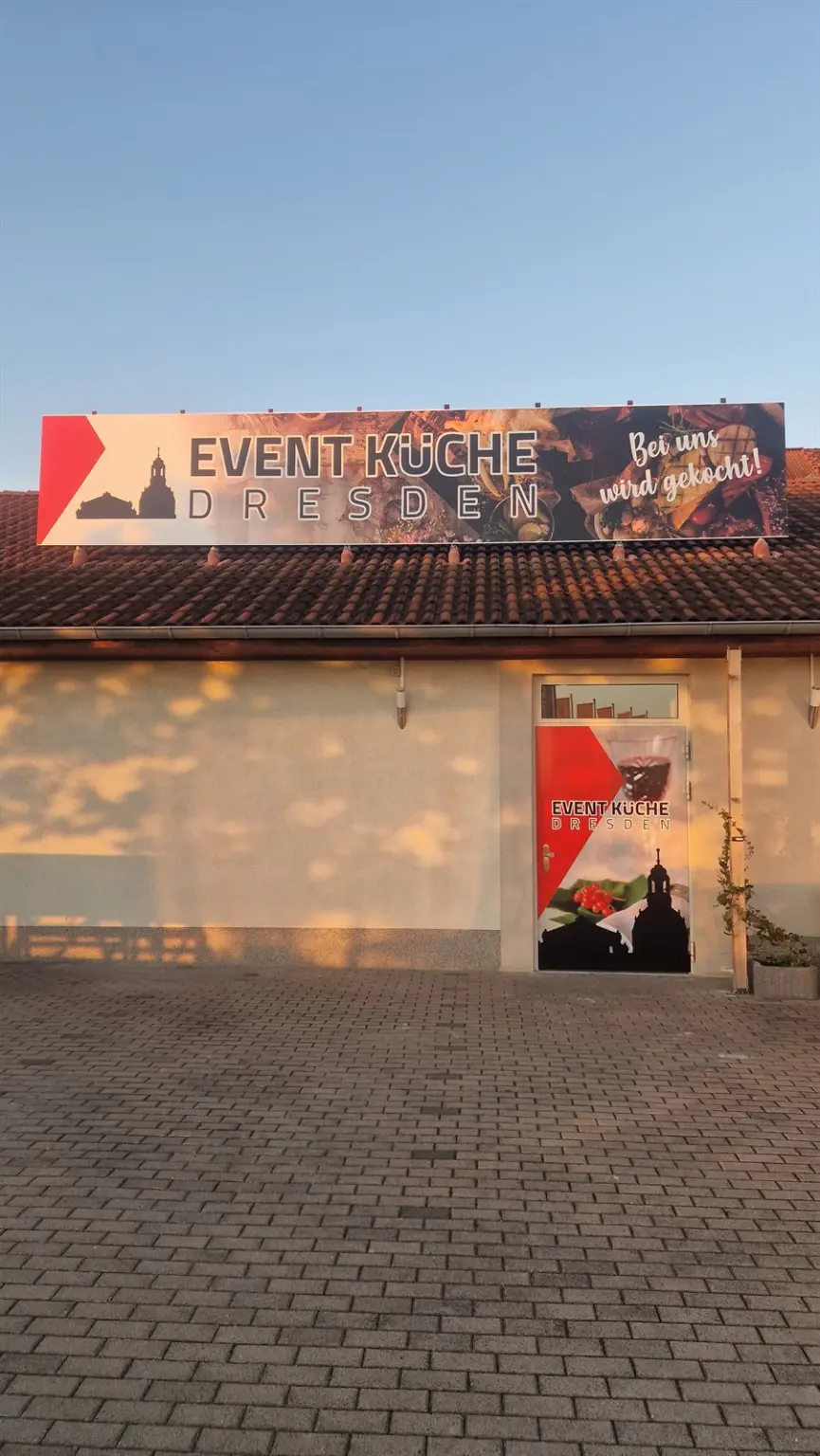Eingangsfront der Event-Küche Dresden im Morgenlicht, Banner auf dem Dach und Türgrafik golden beleuchtet