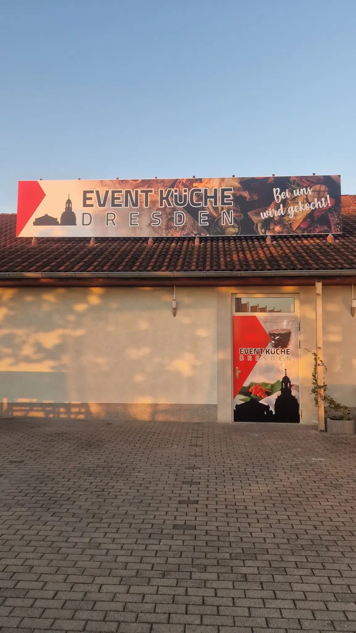 Außenansicht der Event-Küche Dresden im Abendlicht: Werbebanner auf dem Dach, Eingangstür mit Logo, davor der gepflasterte Hof