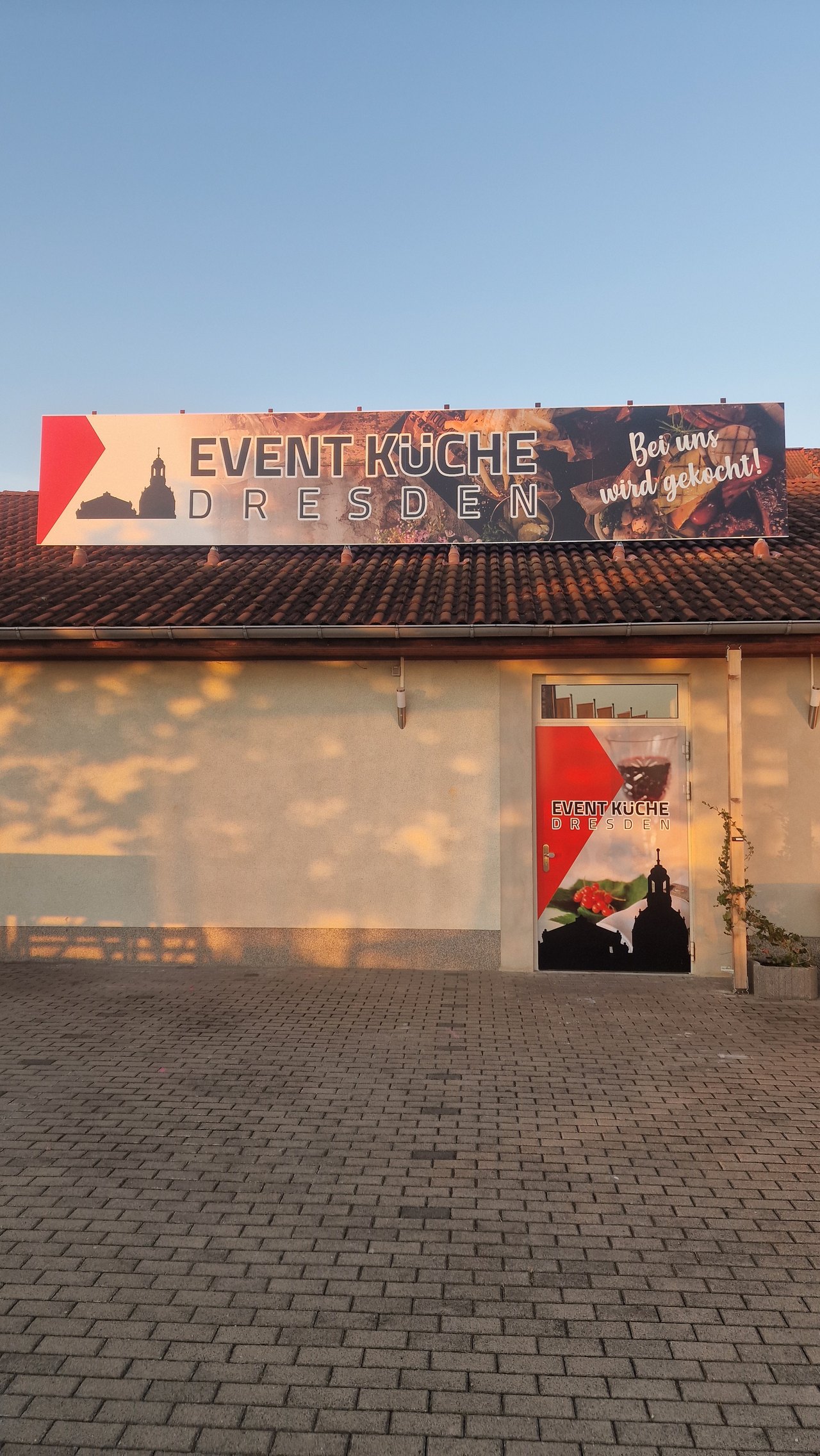 Außenansicht der Event-Küche Dresden im Abendlicht: Werbebanner auf dem Dach, Eingangstür mit Logo, davor der gepflasterte Hof