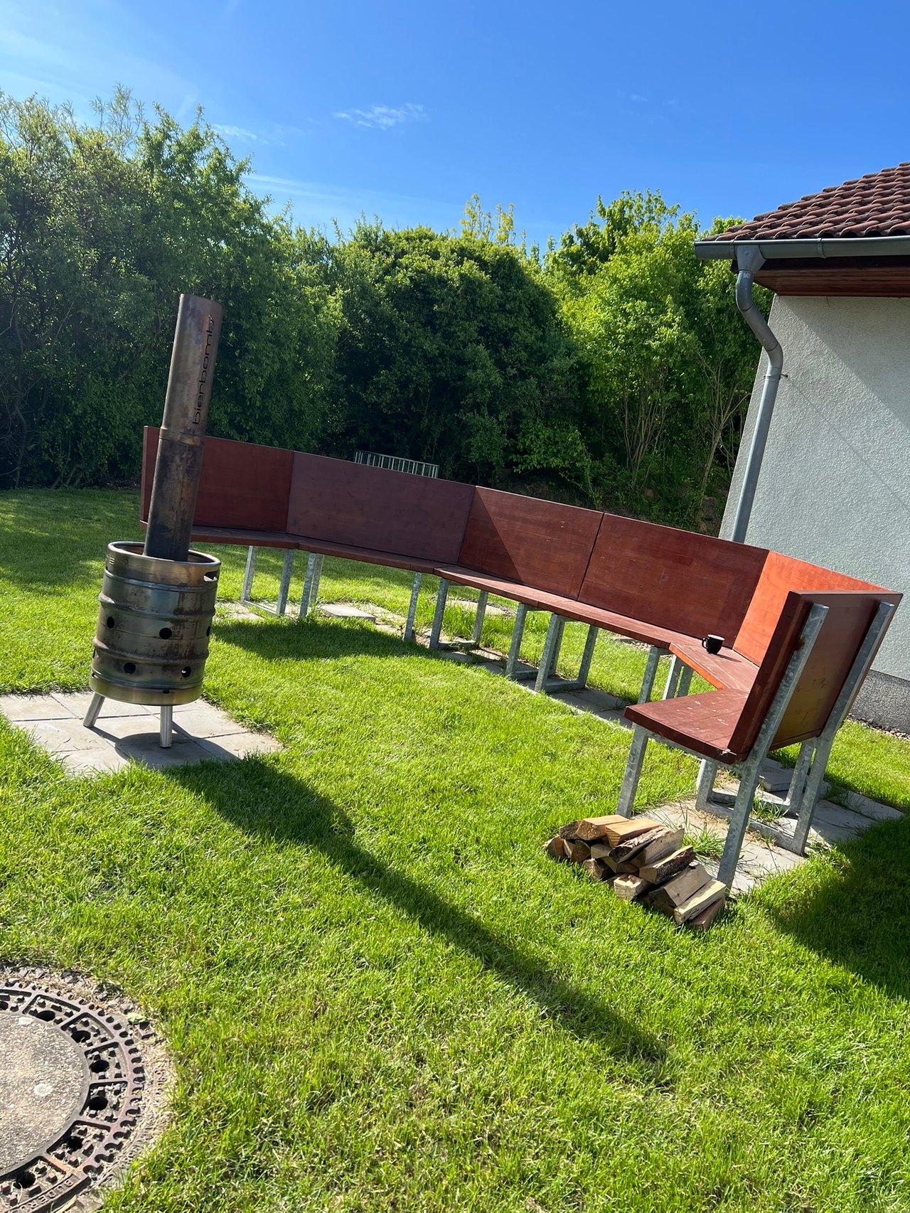Der Außenbereich im Sommer: geschwungene Sitzbank auf dem Rasen, davor die bierbomb Feuertonne und gestapeltes Brennholz