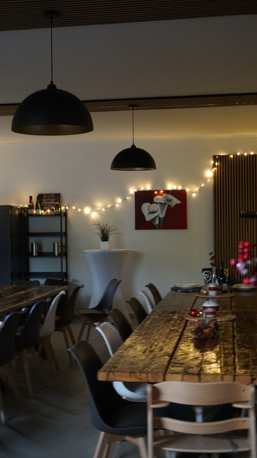 Der Raum abends: zwei Altholztafeln mit grau-weißen Stühlen, ein bezogener Stehtisch und Lichterketten an der Wand