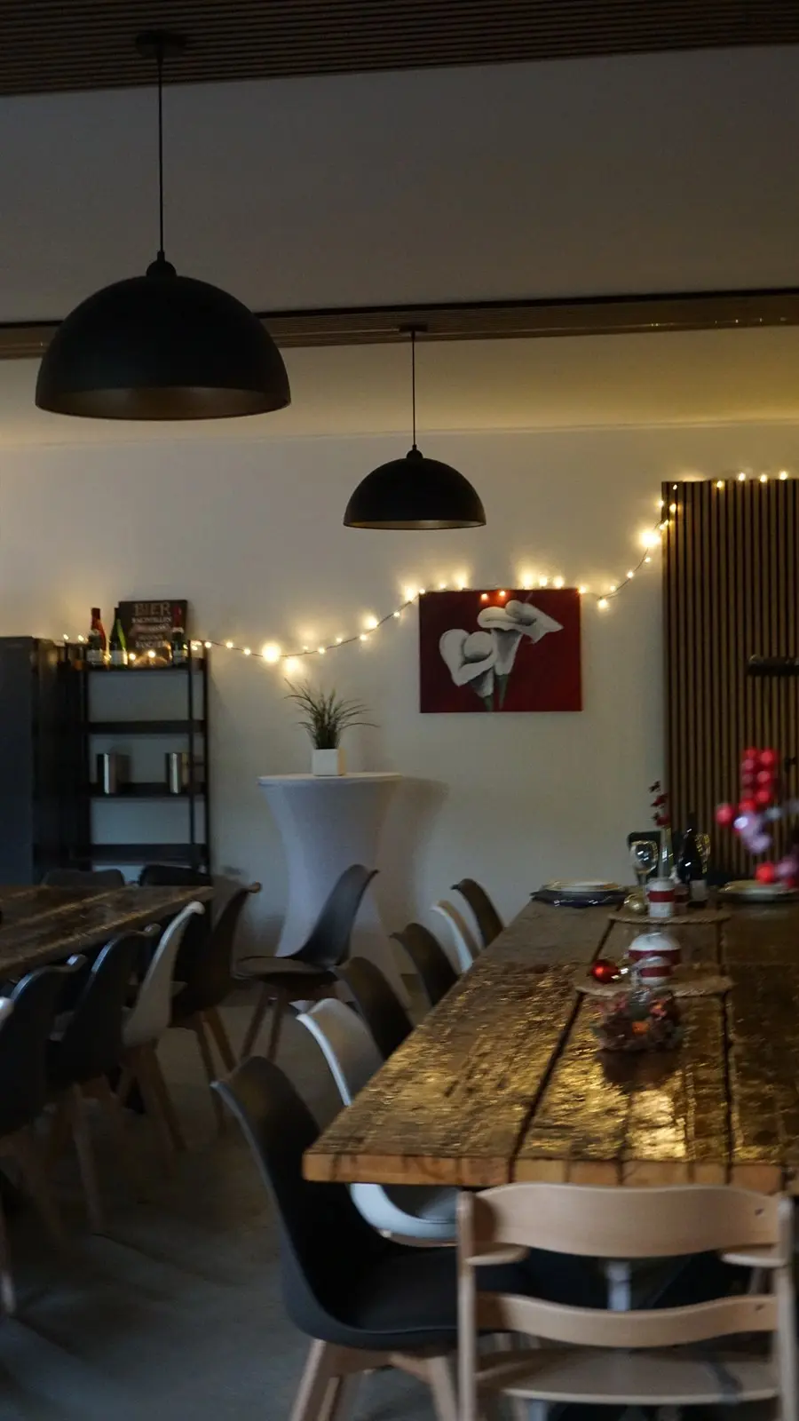 Der Raum abends: zwei Altholztafeln mit grau-weißen Stühlen, ein bezogener Stehtisch und Lichterketten an der Wand