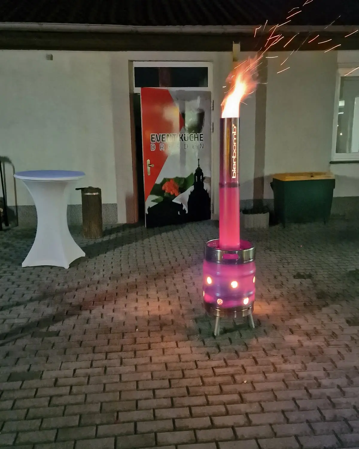 Die bierbomb Feuertonne brennt abends vor dem Eingang der Event-Küche Dresden, daneben ein Stehtisch